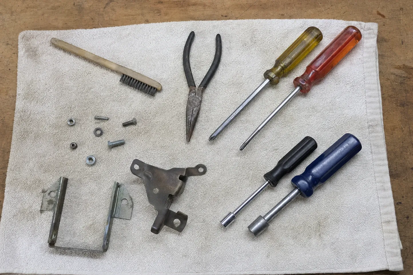 Workbench flat-lay of a wire brush, pliers, screwdrivers, nut drivers, two sheet-metal brackets and loose hardware