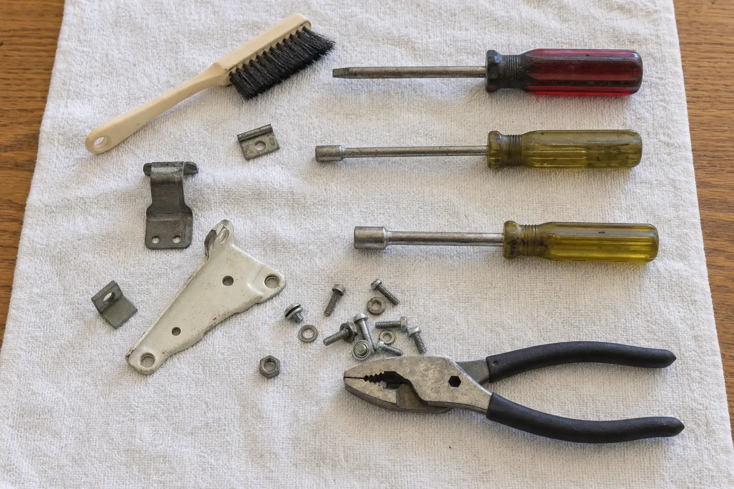 Stiff-bristled brush beside a red-handled screwdriver, two yellow nut drivers, slip-joint pliers, two steel brackets, keeper clips and an assortment of screws and nuts